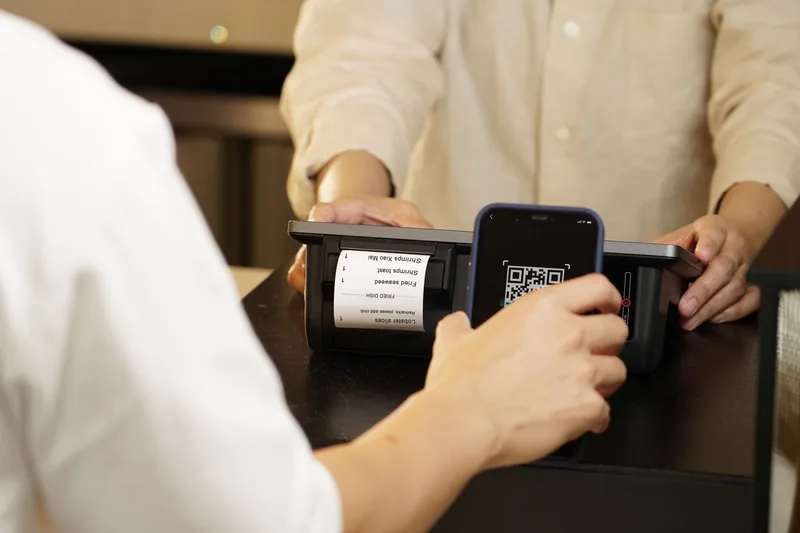 Customer holding a phone with a QR code to a payment terminal that prints a receipt at a shop counter